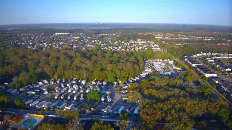 Aerial view of an RV campground nestled among trees, with residential neighborhoods and a distant skyline.