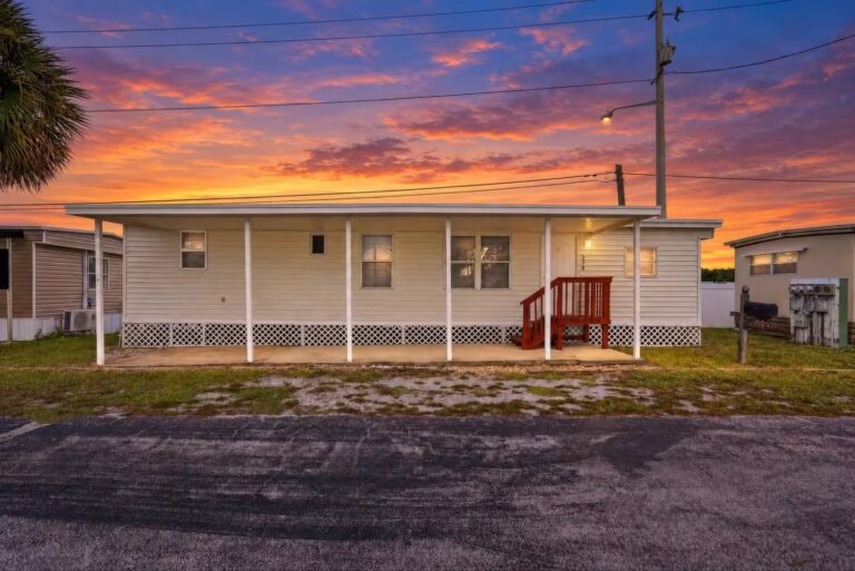 Single-wide mobile home with covered porch and red stairs at sunset.