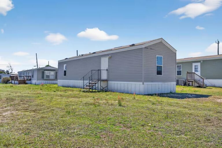 Gray mobile home with white skirting and stairs, surrounded by grass and other manufactured homes under blue sky.