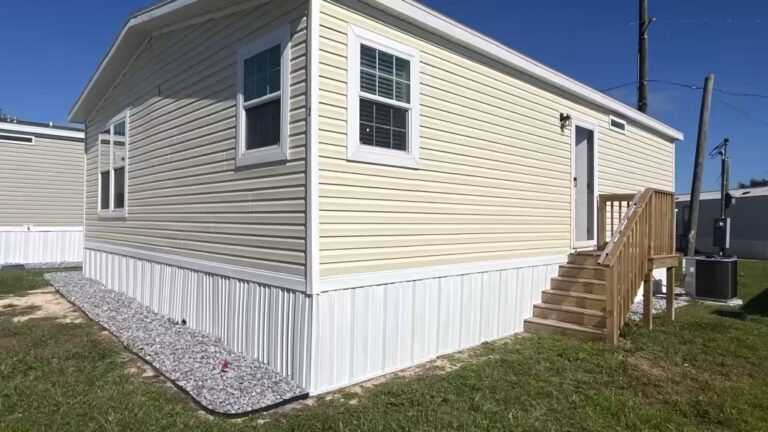 Yellow single-wide mobile home with white trim, wooden stairs, and gravel drainage bed under blue sky.