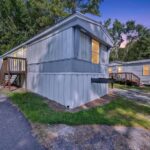 Gray mobile home with wooden deck stairs at dusk, trees and white pickup truck in background.