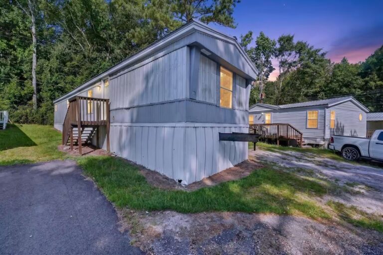 Gray mobile home with wooden deck stairs at dusk, trees and white pickup truck in background.