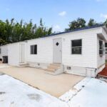 White single-wide mobile home with blue roof, concrete patio, steps, and trees in background.