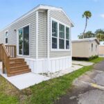 Small mobile home with sage green siding, wooden stairs, and deck on a grassy lot under blue sky.