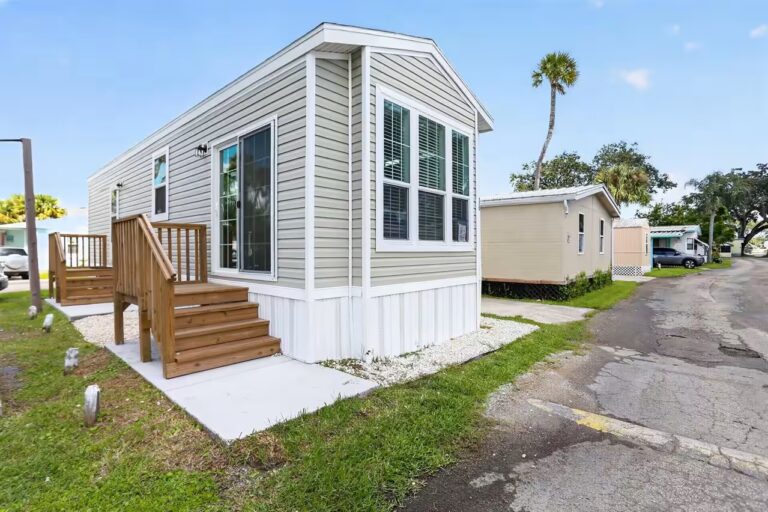 Small mobile home with sage green siding, wooden stairs, and deck on a grassy lot under blue sky.