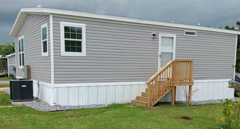 Gray single-wide mobile home with white trim, wooden porch stairs, and HVAC unit under cloudy sky.