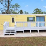 Yellow single-wide mobile home with white deck and railing, green shutters, and blue curtains.