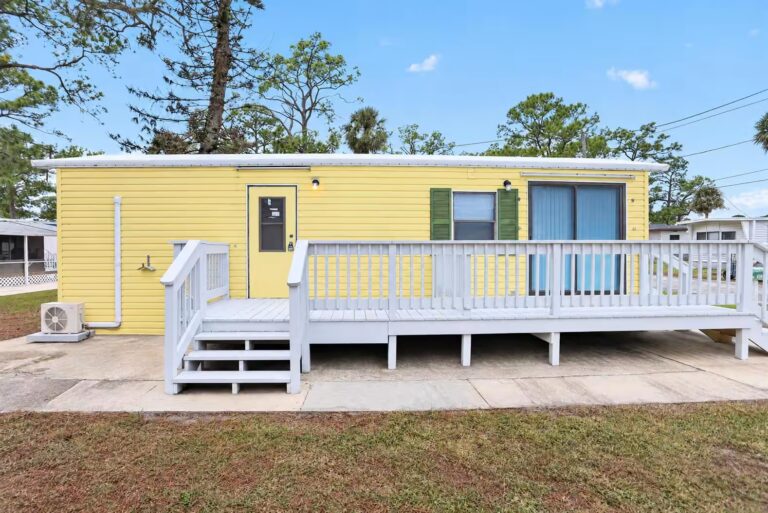 Yellow single-wide mobile home with white deck and railing, green shutters, and blue curtains.
