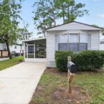 White mobile home with screened porch, concrete driveway, mailbox, and green shrubs under blue sky.