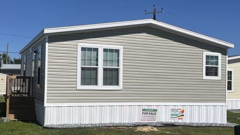 Beige manufactured home with "FOR SALE" sign under clear blue sky.