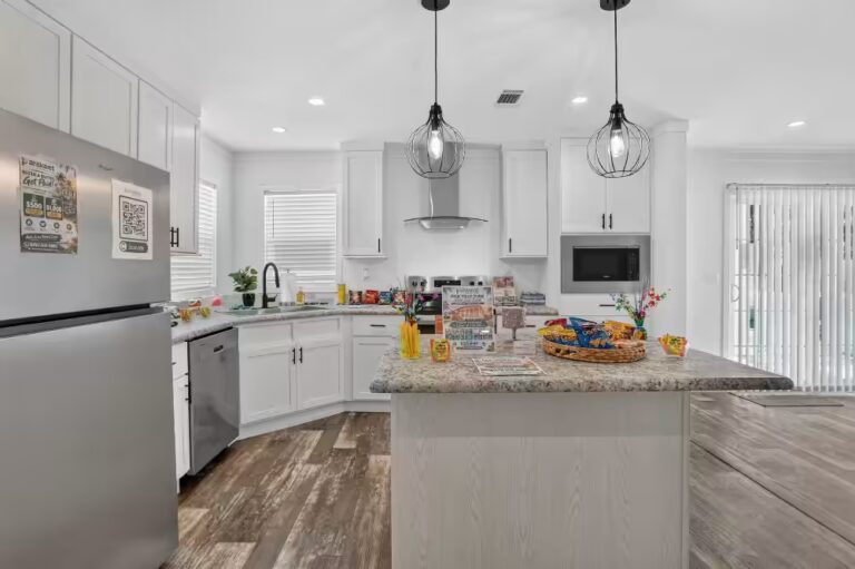 White kitchen with granite island, caged pendant lights, and stainless steel appliances.