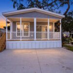 Gray mobile home with white porch and wooden stairs at dusk, concrete driveway in foreground