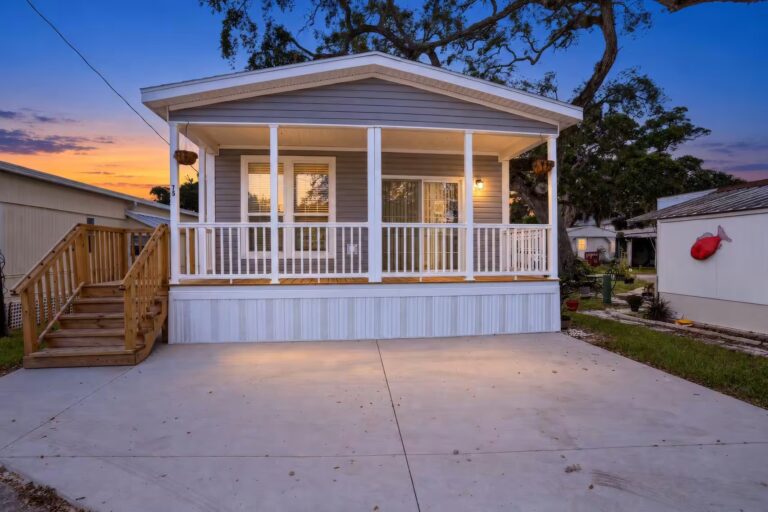 Gray mobile home with white porch and wooden stairs at dusk, concrete driveway in foreground