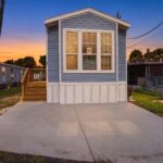 Blue mobile home with For Sale sign in window, wooden stairs and concrete driveway at dusk