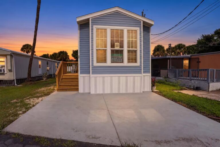 Blue mobile home with For Sale sign in window, wooden stairs and concrete driveway at dusk