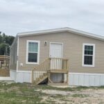 Beige mobile home with white front door and wooden porch stairs under an overcast sky.