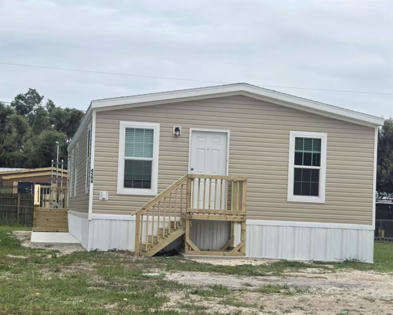 Beige mobile home with white front door and wooden porch stairs under an overcast sky.