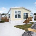 Yellow mobile home with wooden deck stairs, "for sale" sign in window, clear blue sky.