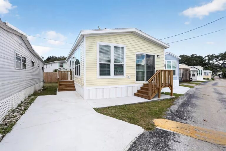 Yellow mobile home with wooden deck stairs, "for sale" sign in window, clear blue sky.