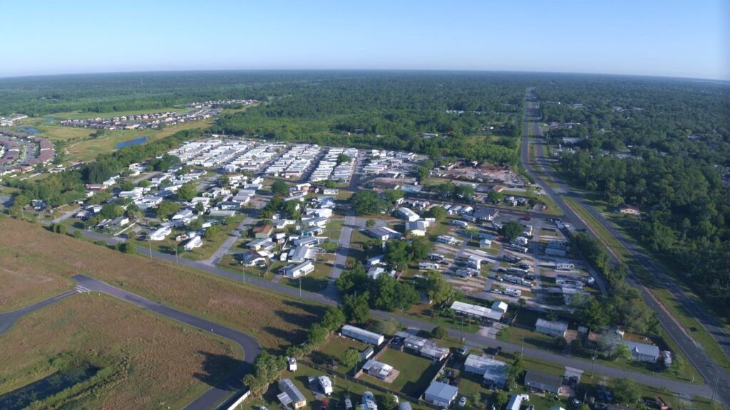 Aerial view of a mobile home park with rows of white units, bordered by trees, fields, and a highway.