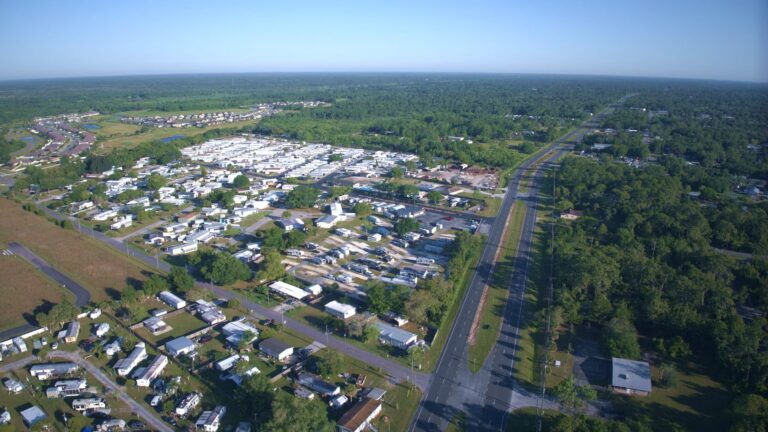 Aerial view of a mobile home park with white trailers beside a road, surrounded by trees.