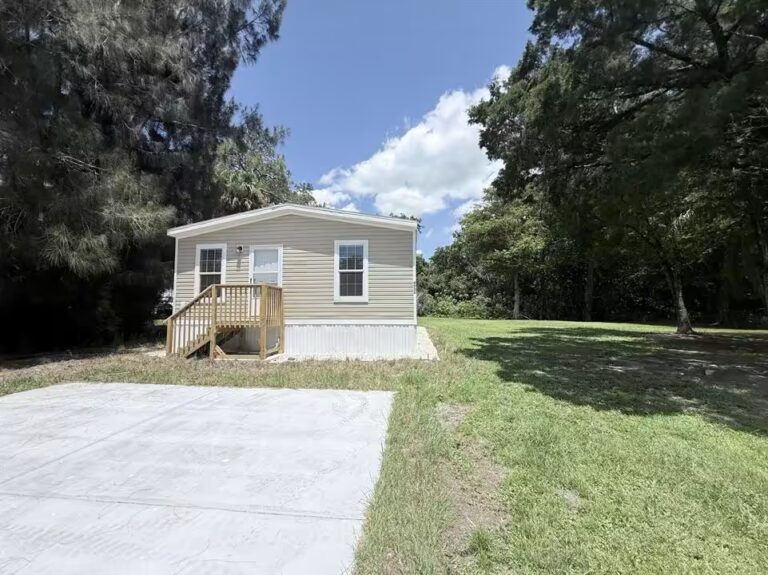 Small beige single-story mobile home with wooden ramp, surrounded by trees and grass under a blue sky.