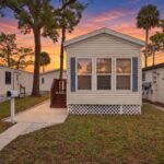 Cream mobile home with blue shutters and lattice skirting beneath a colorful sunset sky with palm trees.