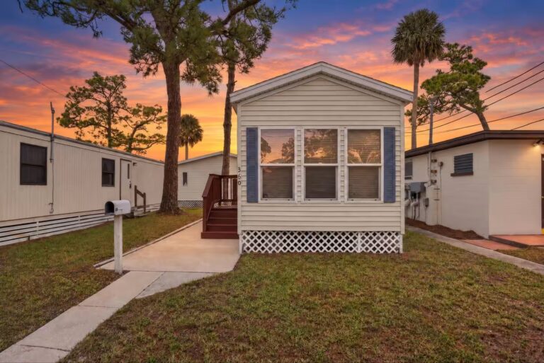 Cream mobile home with blue shutters and lattice skirting beneath a colorful sunset sky with palm trees.