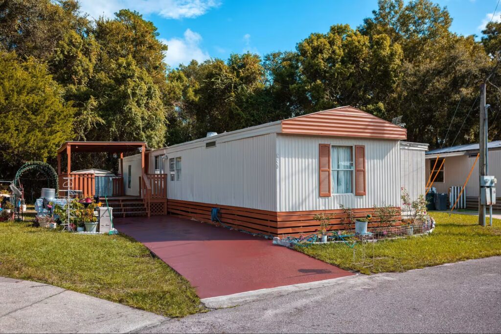 White mobile home with red trim and shutters, red driveway, wooden deck, and green trees behind.
