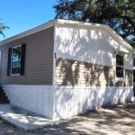 Mobile home with white and tan siding surrounded by large oak trees under a clear blue sky.