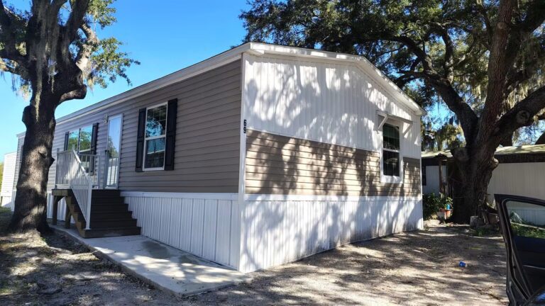 Mobile home with white and tan siding surrounded by large oak trees under a clear blue sky.