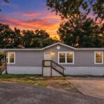 Gray mobile home with porch steps beneath a vibrant orange and purple sunset sky, framed by trees.
