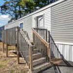 Wooden stairs and railing lead up to the door of a white mobile home with vinyl siding.