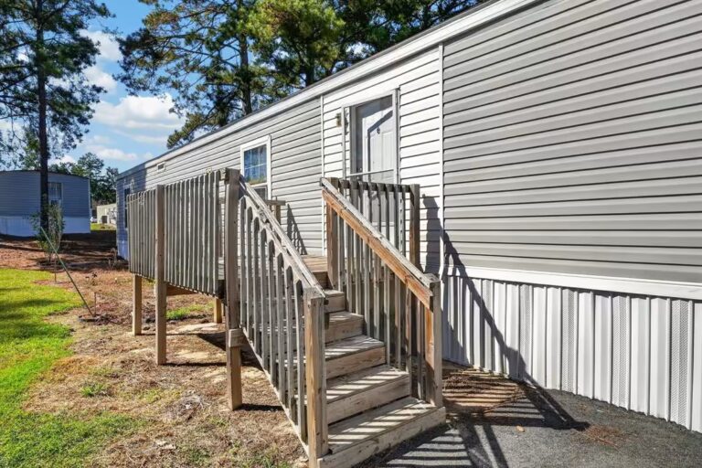 Wooden stairs and railing lead up to the door of a white mobile home with vinyl siding.