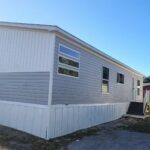 Gray and white mobile home with stairs, palm trees, and red shed under a blue sky.