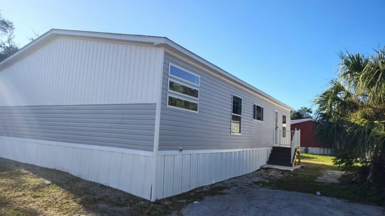Gray and white mobile home with stairs, palm trees, and red shed under a blue sky.