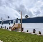 Single-wide mobile home with white and blue siding, wooden stairs, shrubs, and palm trees under cloudy sky
