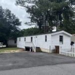 White mobile home with FOR SALE sign in window, wooden stairs, white picket fence, tall trees behind.