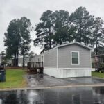 Gray mobile home with wooden deck and stairs on a wet driveway under an overcast sky, tall pines behind.