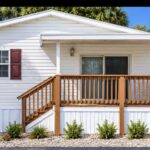 White mobile home with wooden porch, stairs, and red shutters, surrounded by palm trees and landscaping.