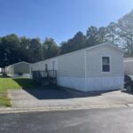 White single-wide mobile home with dark roof, driveway, and trees under a clear blue sky.