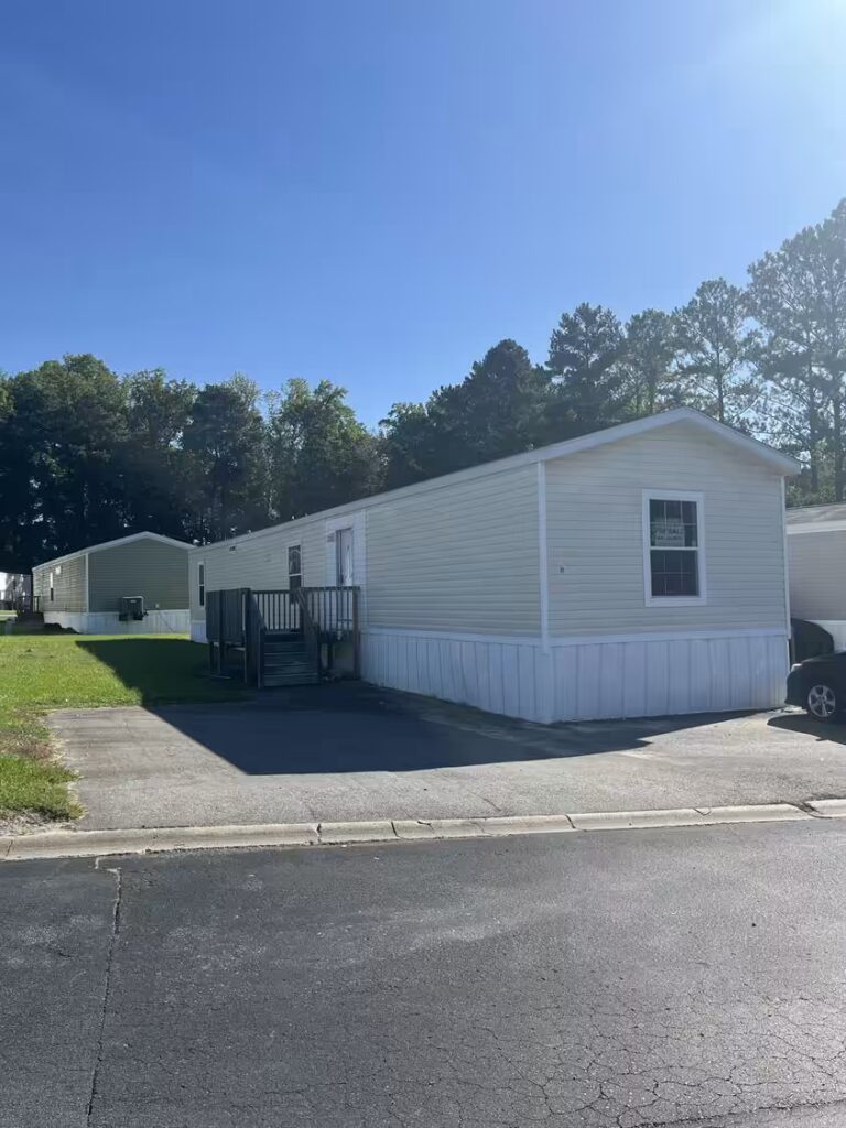White single-wide mobile home with dark roof, driveway, and trees under a clear blue sky.